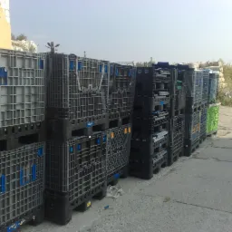 Rząd ułożonych skrzyniopalet składanych z tworzywa sztucznego, niektóre w kolorze szarym, inne czarne i jeden zielony, stojących na betonowym podłożu, z widocznym fragmentem budynku w tle.